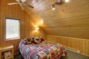 Een slaapkamer met een houten plafond, een bed met een quilt met patroon, een nachtkastje, een raam met jaloezieën en een plafondventilator.