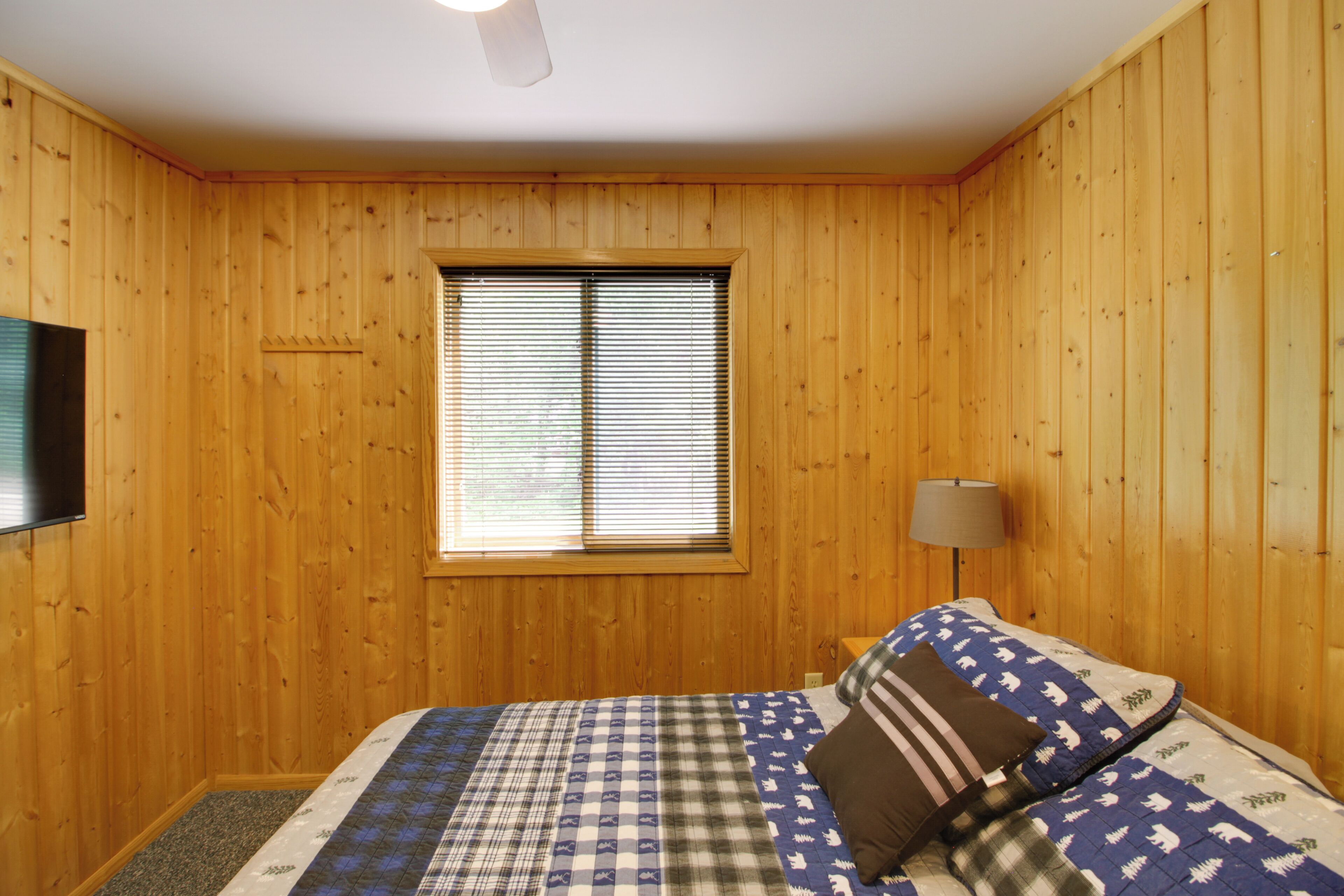 Quarto com paredes de madeira, uma cama com roupa de cama estampada, uma janela com persianas, um abajur e um ventilador de teto.