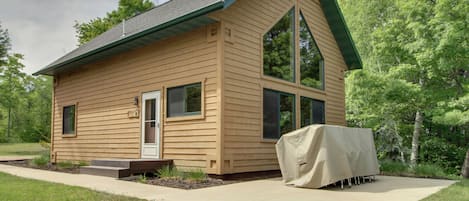 Une petite cabane en bois, d’un seul étage, avec un toit à pignon, un espace extérieur couvert aménagé en coin salon et une porte d’entrée.