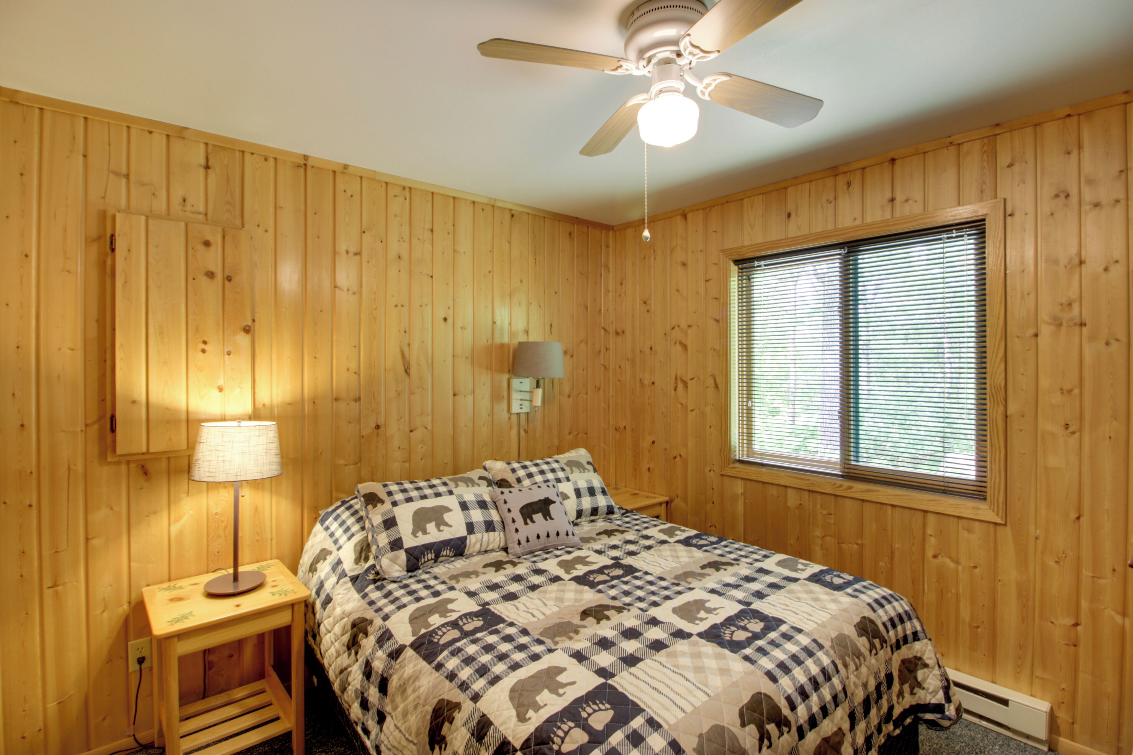A room with wooden walls, a ceiling fan, a bed with a checkered quilt, a bedside lamp, and a window with blinds.