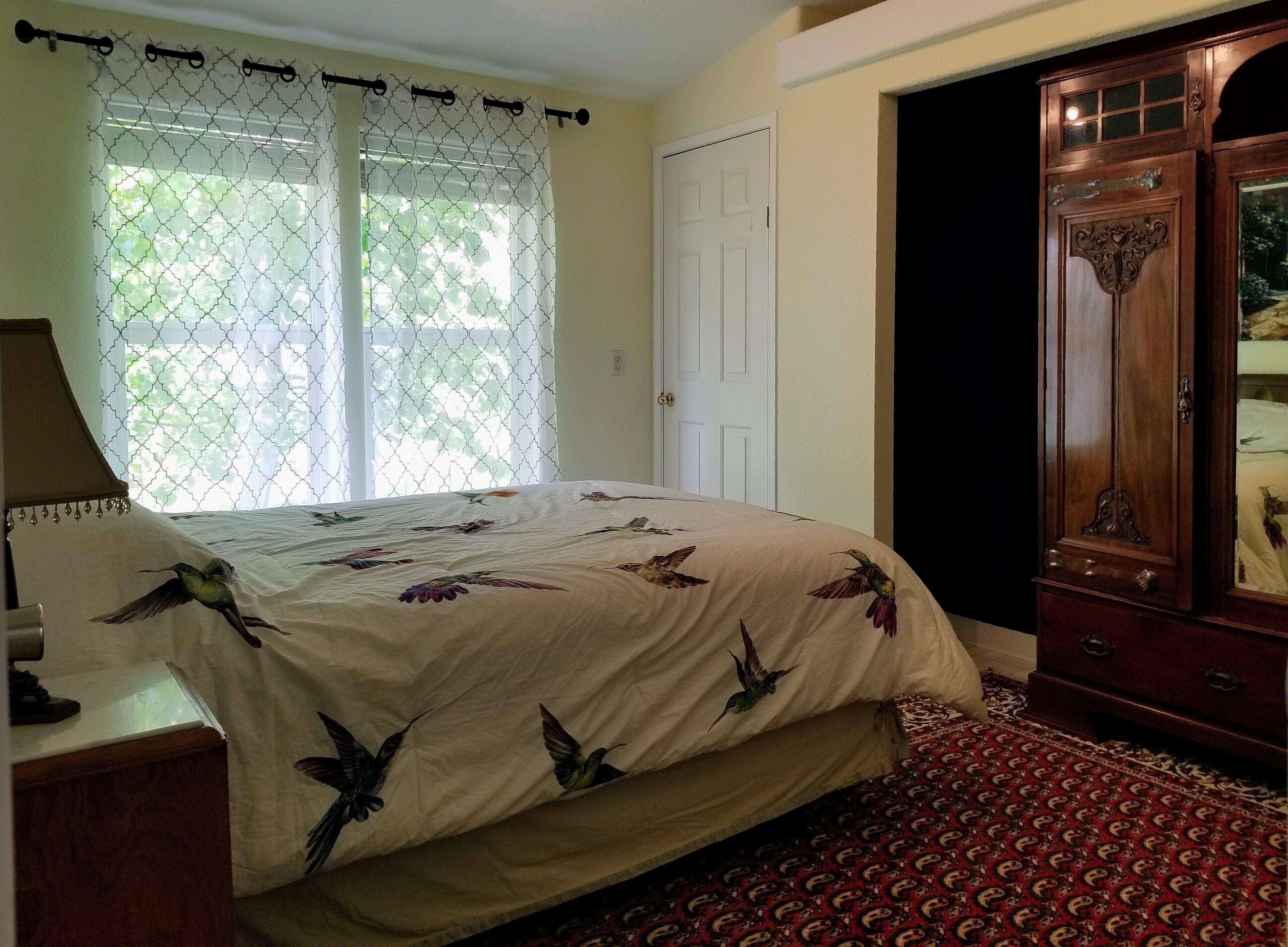 A bedroom with a bed, a wooden wardrobe, a window with a white curtain, and a patterned carpet.
