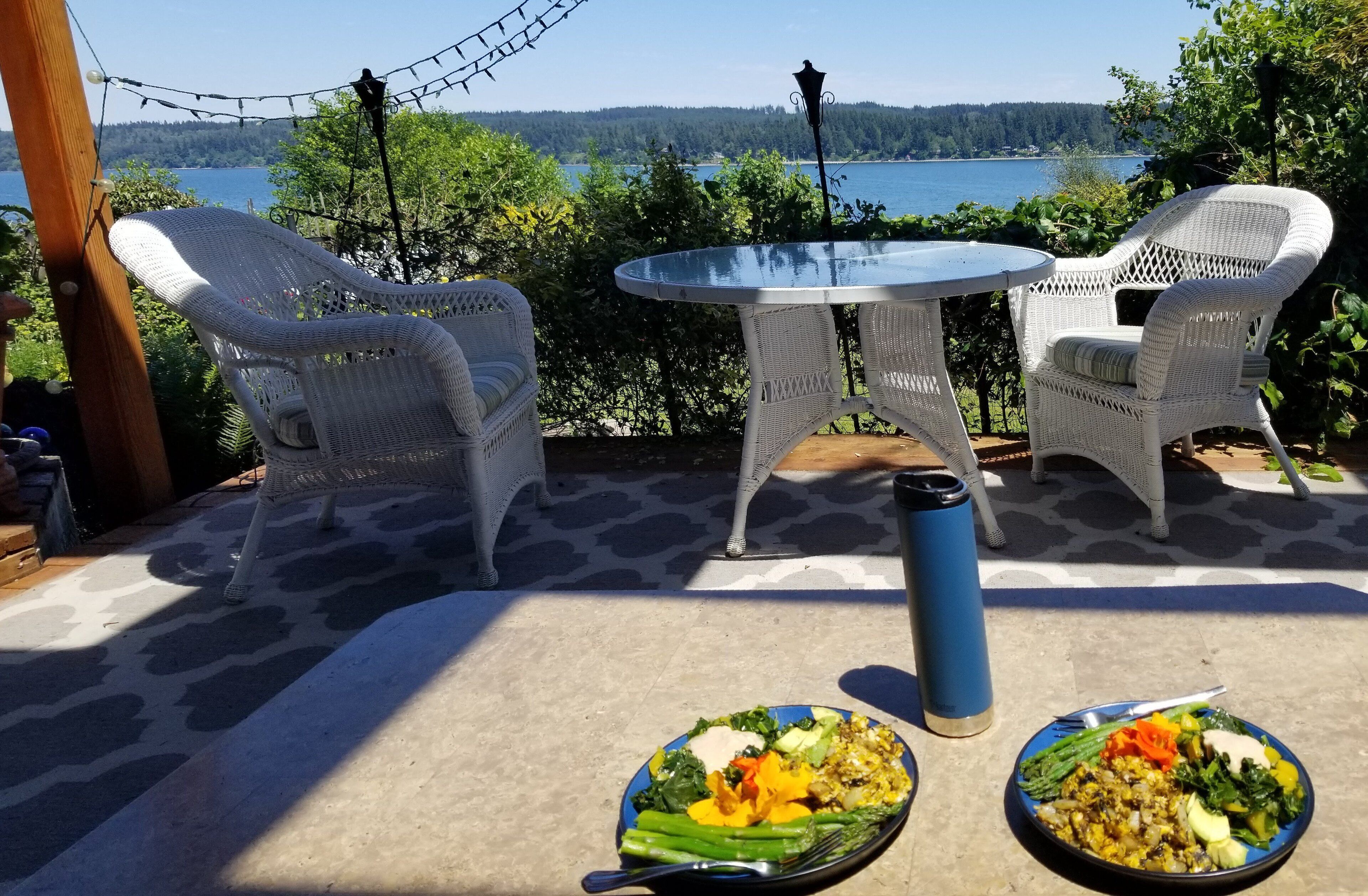 En patio med to fletstole og et rundt bord, med udsigt over en sø med skovkant.