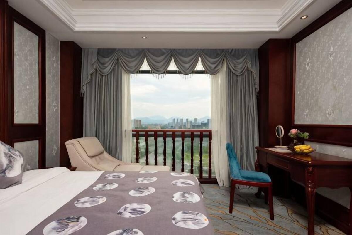Ein Hotelzimmer mit einem großen Bett, einem Schreibtisch mit Stuhl und Blick auf die Stadt durch einen Balkon, sowie einem Stadtbild durch das Fenster.