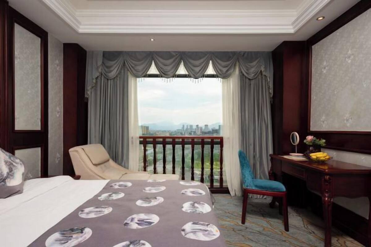 Ein Hotelzimmer mit einem Bett, zwei Stühlen, einem Schreibtisch und Blick auf eine Stadtlandschaft durch große Fenster.