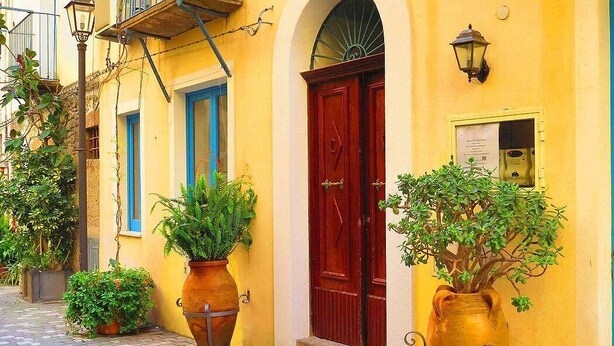 Une rue étroite bordée d’immeubles jaunes, avec une porte rouge et des plantes en pot.