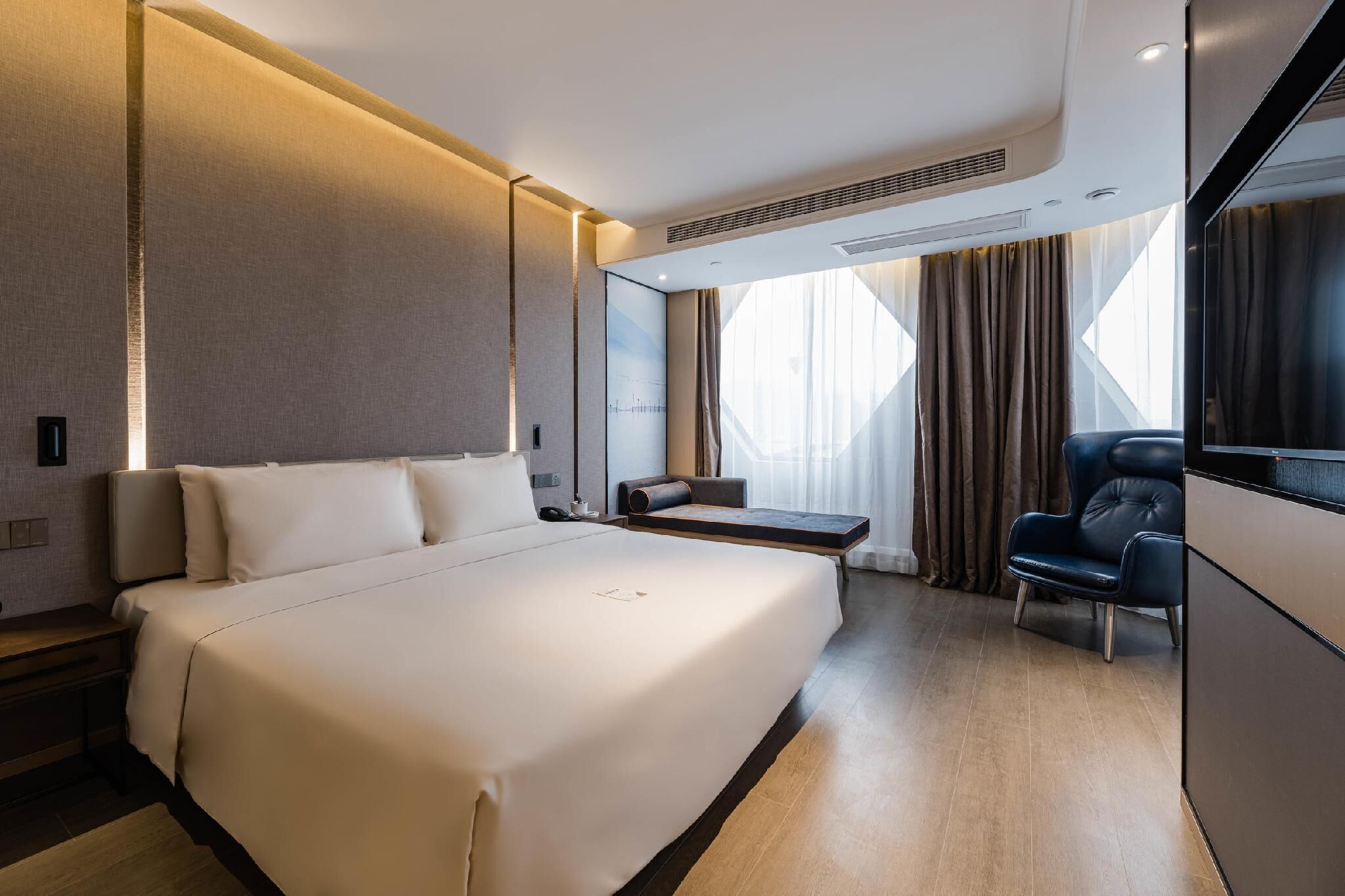 Quarto de hotel moderno com uma cama grande, área de estar, escrivaninha e vista da cidade através de janelas amplas.