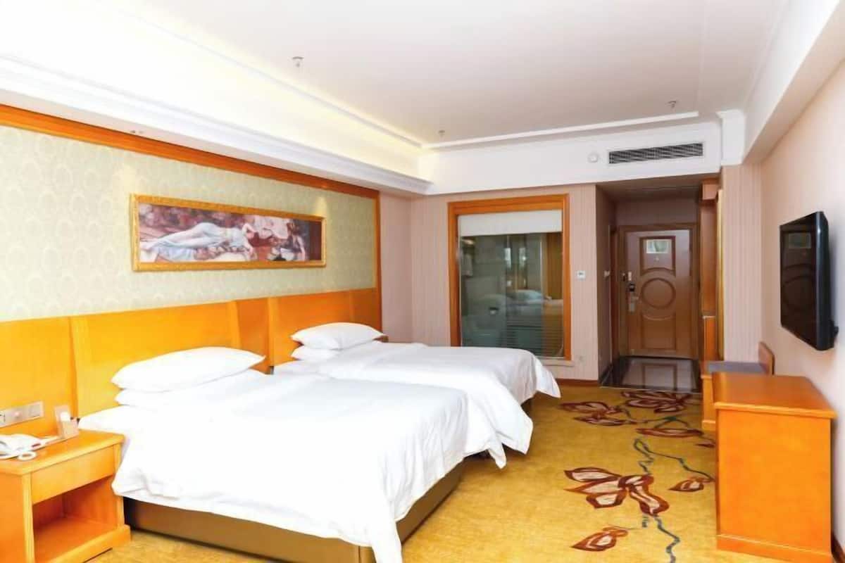 A hotel room with two beds, a painting on the wall, a television, and a carpet with a leaf pattern.