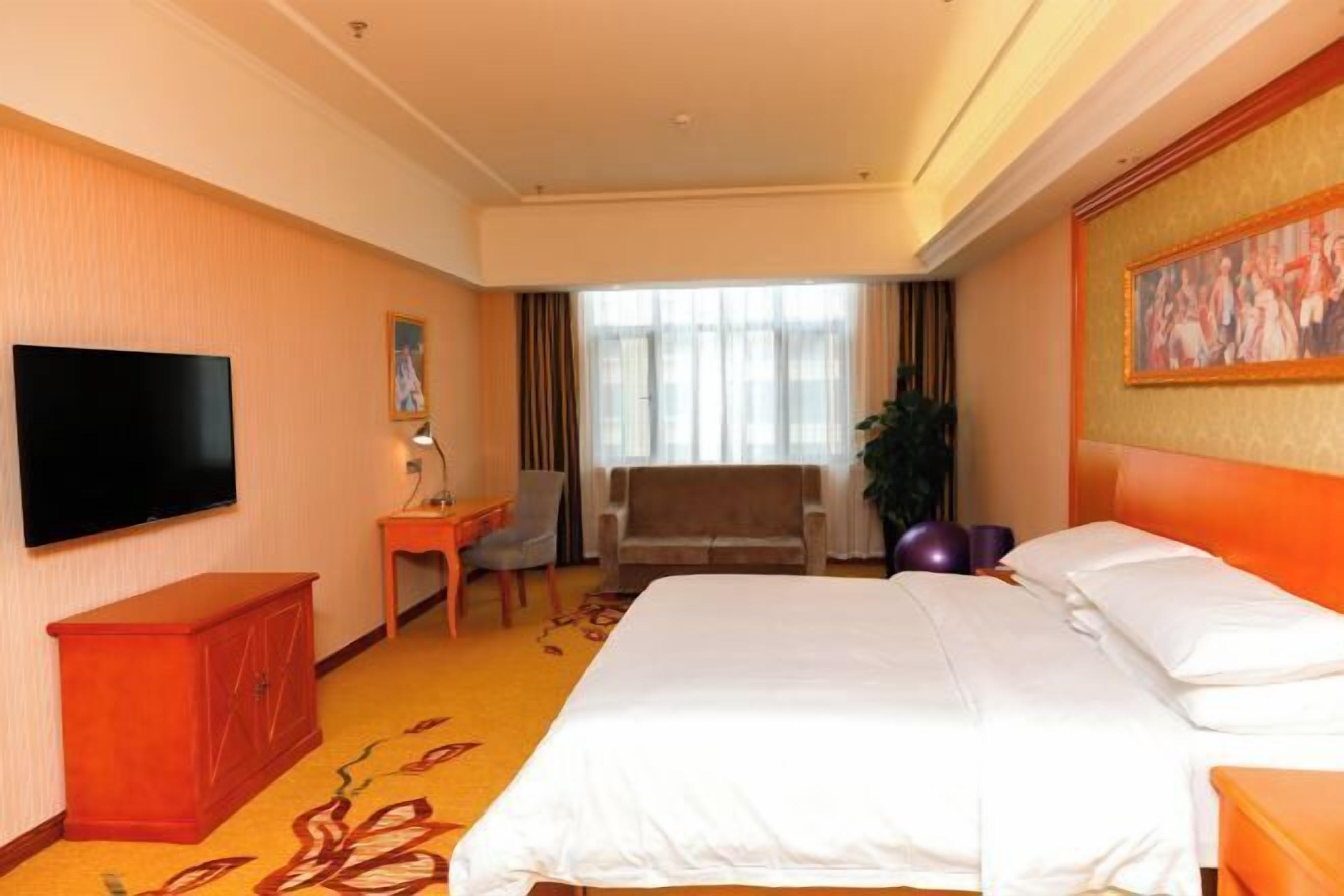 A hotel room with a bed, a TV, a desk, a chair, and a window with curtains.