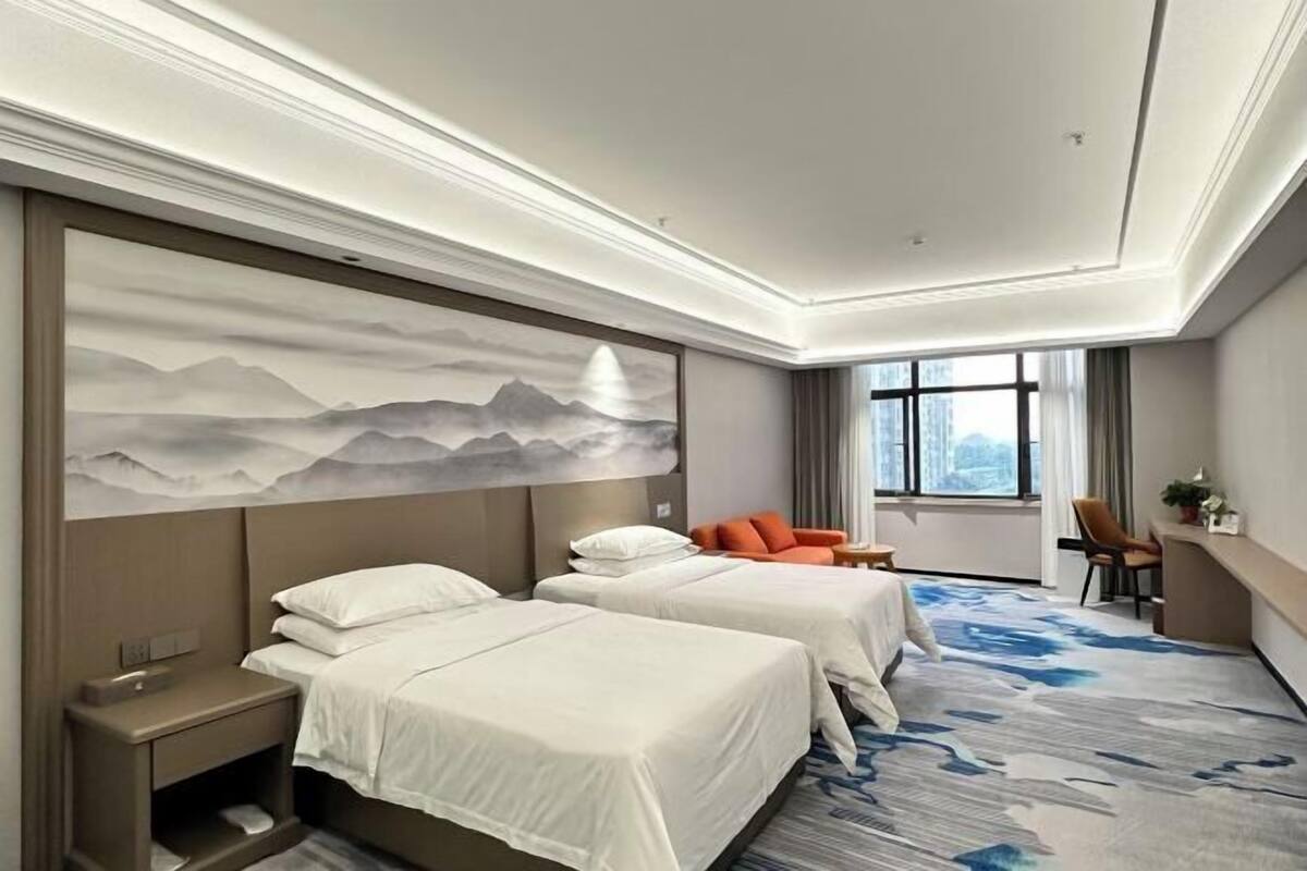 A hotel room with two beds, a large wall painting, a desk, and a view of the city through the window.