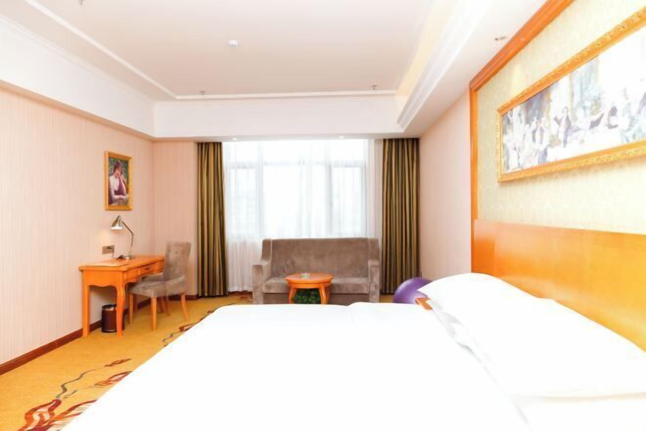 A hotel room with a bed, a desk, a chair, a small table, and a large window with curtains.