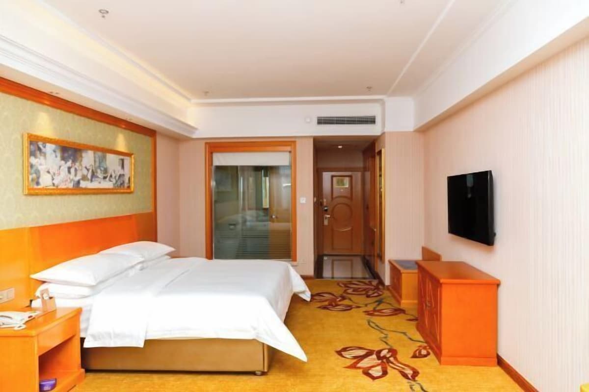 Habitación de hotel con cama, televisión, un cuadro en la pared y piso alfombrado.