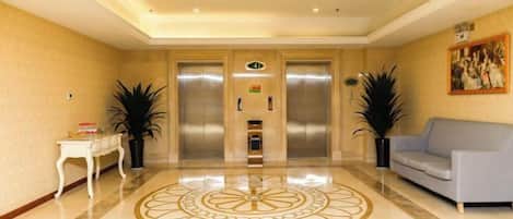 A lobby with a polished marble floor, two elevators, a white console table, a gray sofa, and potted plants.
