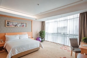 Une chambre d’hôtel avec un grand lit, un bureau, une chaise, une plante en pot et une grande fenêtre avec des rideaux.