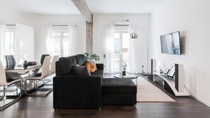A modern living room with a black sofa, a dining table with chairs, a TV, and a large window with curtains.