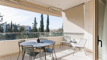 Un balcon avec une table ronde et des chaises, un pot de fleurs et une vue sur les arbres et les bâtiments.