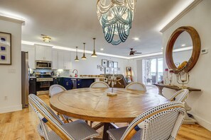 Un coin repas avec une table ronde en bois, des chaises en osier, des luminaires suspendus, un ventilateur de plafond et une cuisine en arrière-plan.