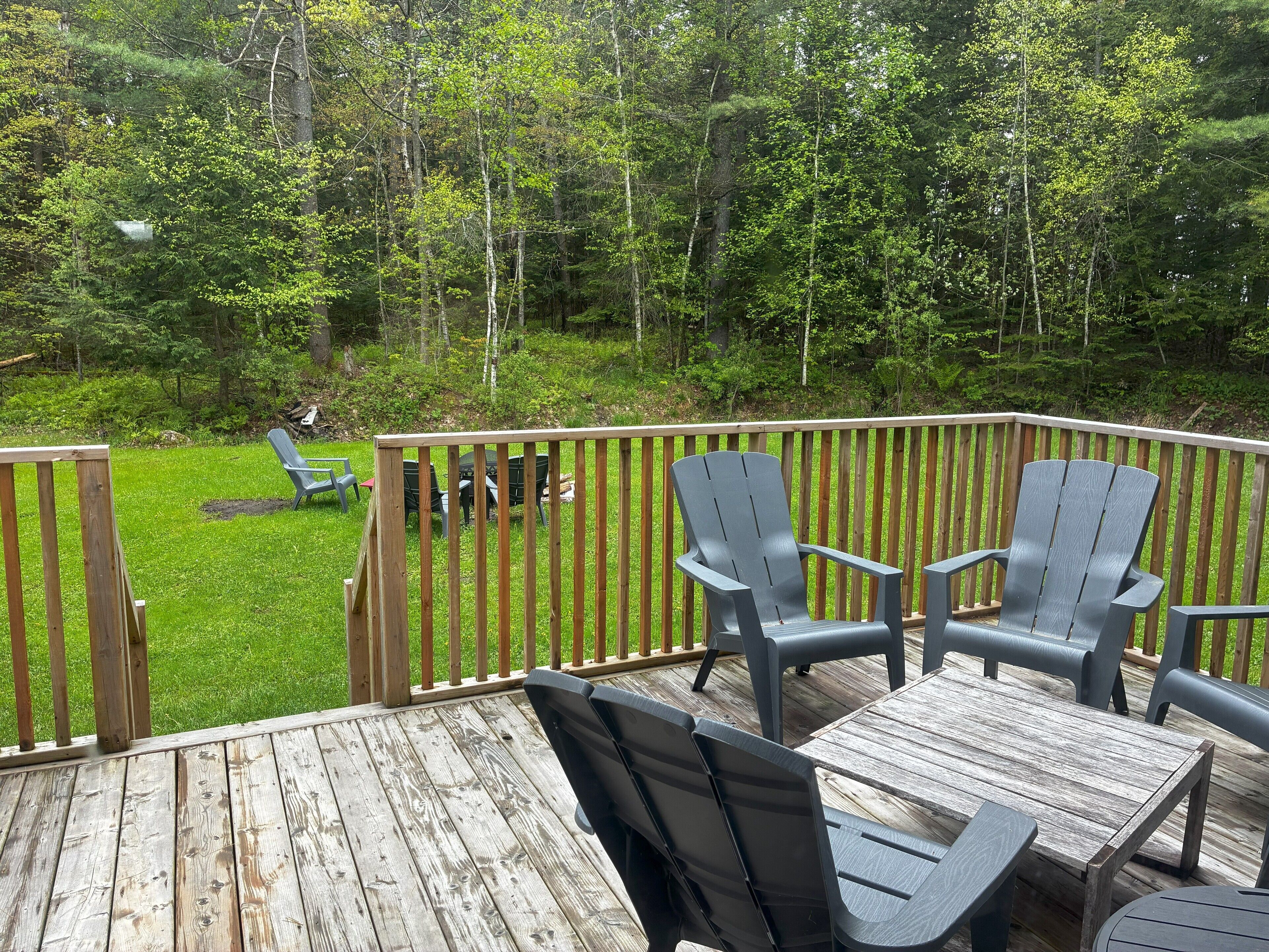 A wooden deck with outdoor furniture, including chairs and a table, overlooking a grassy area with trees.