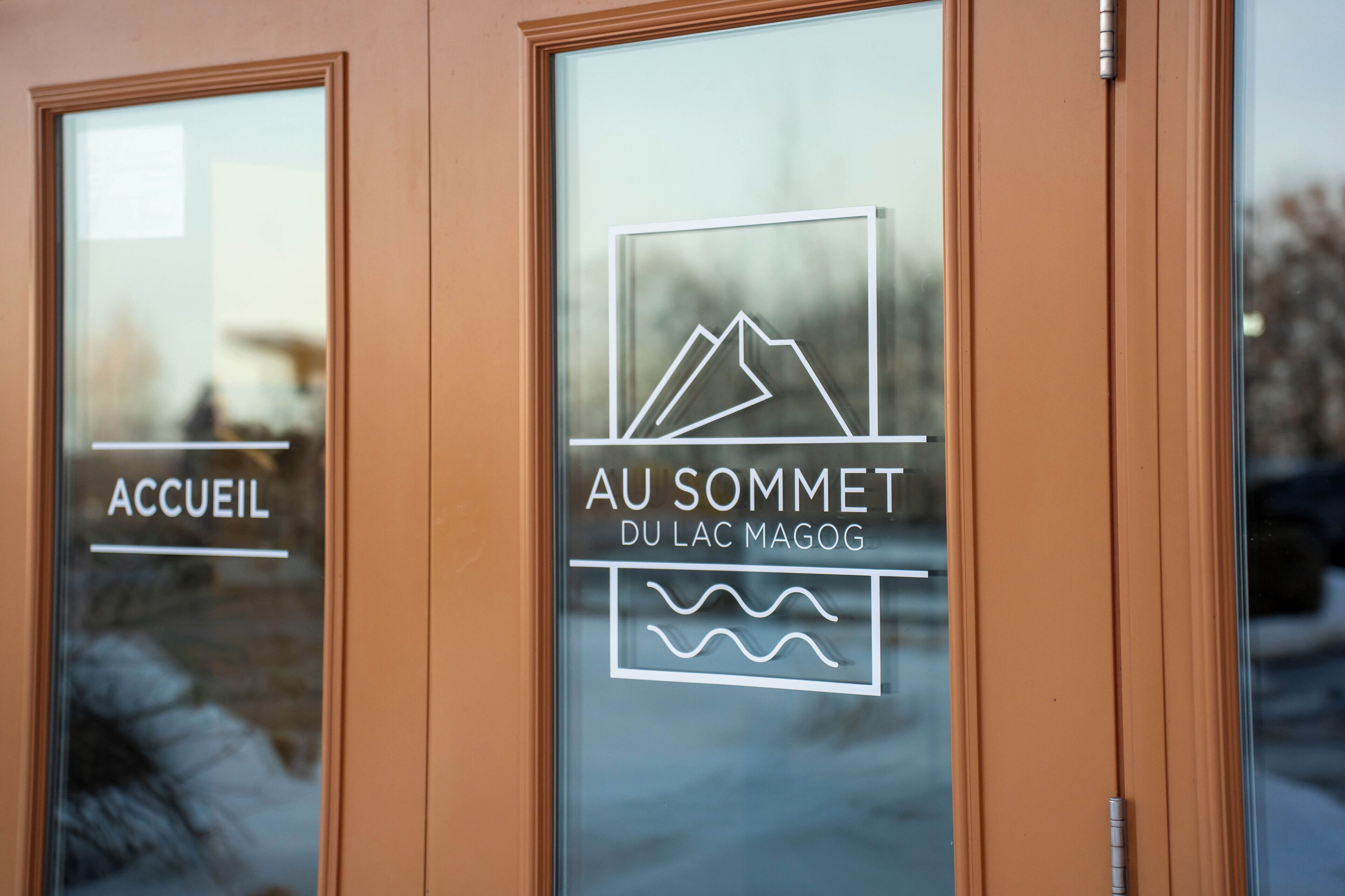 A hotel entrance with glass doors displaying the sign "AU SOMMET DU LAC MAGOG".