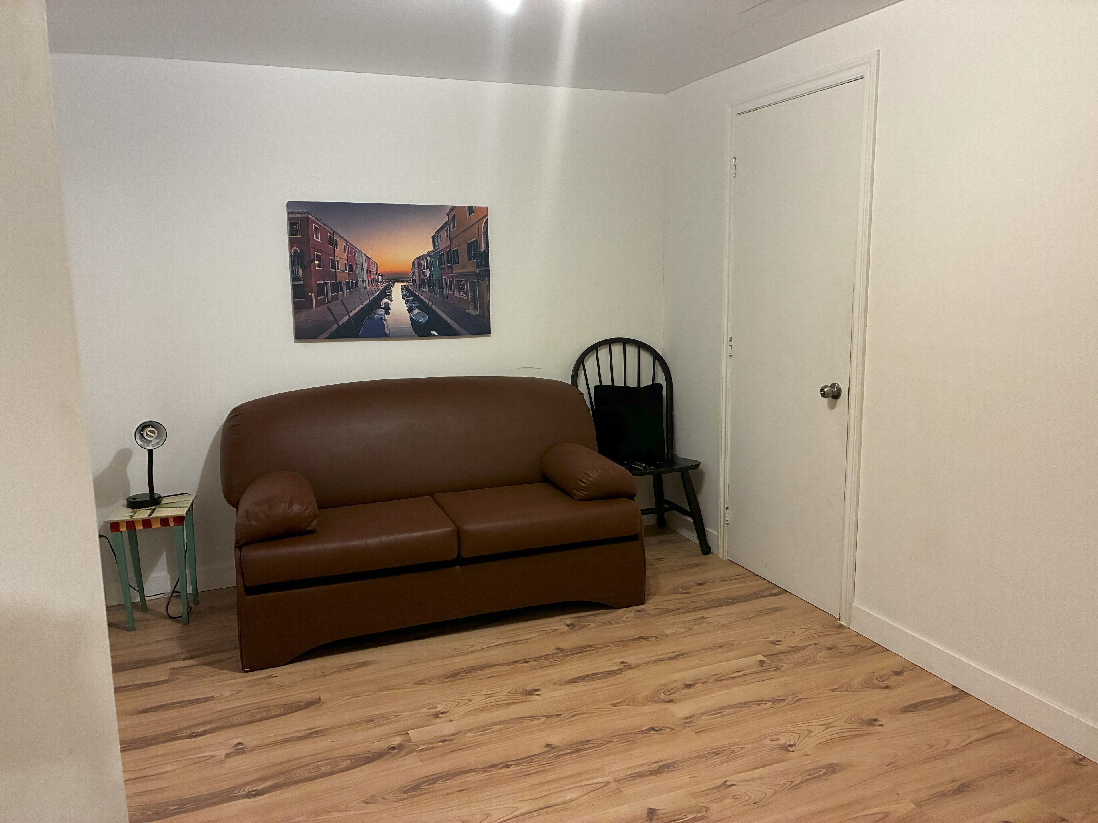 Una habitación con un sofá de cuero marrón, una mesita con una lámpara, una silla y una puerta.