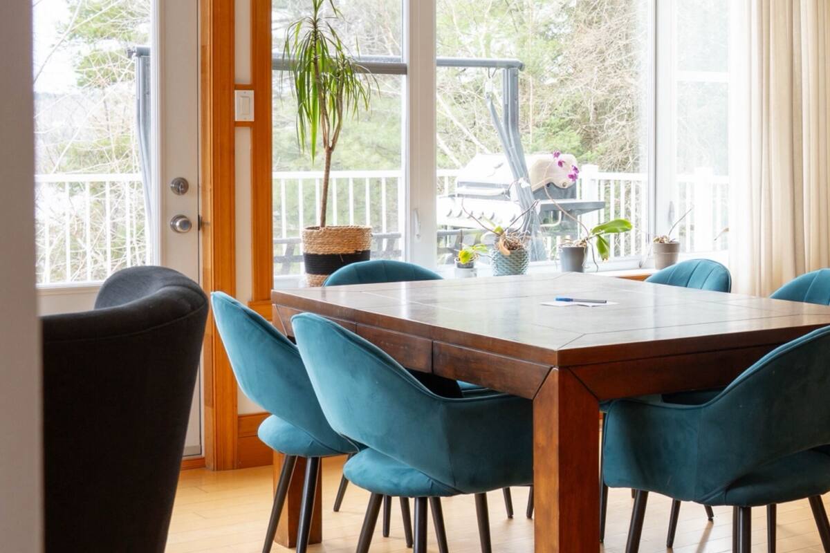 A dining area with a wooden table and blue chairs, large windows, and a view of trees outside.