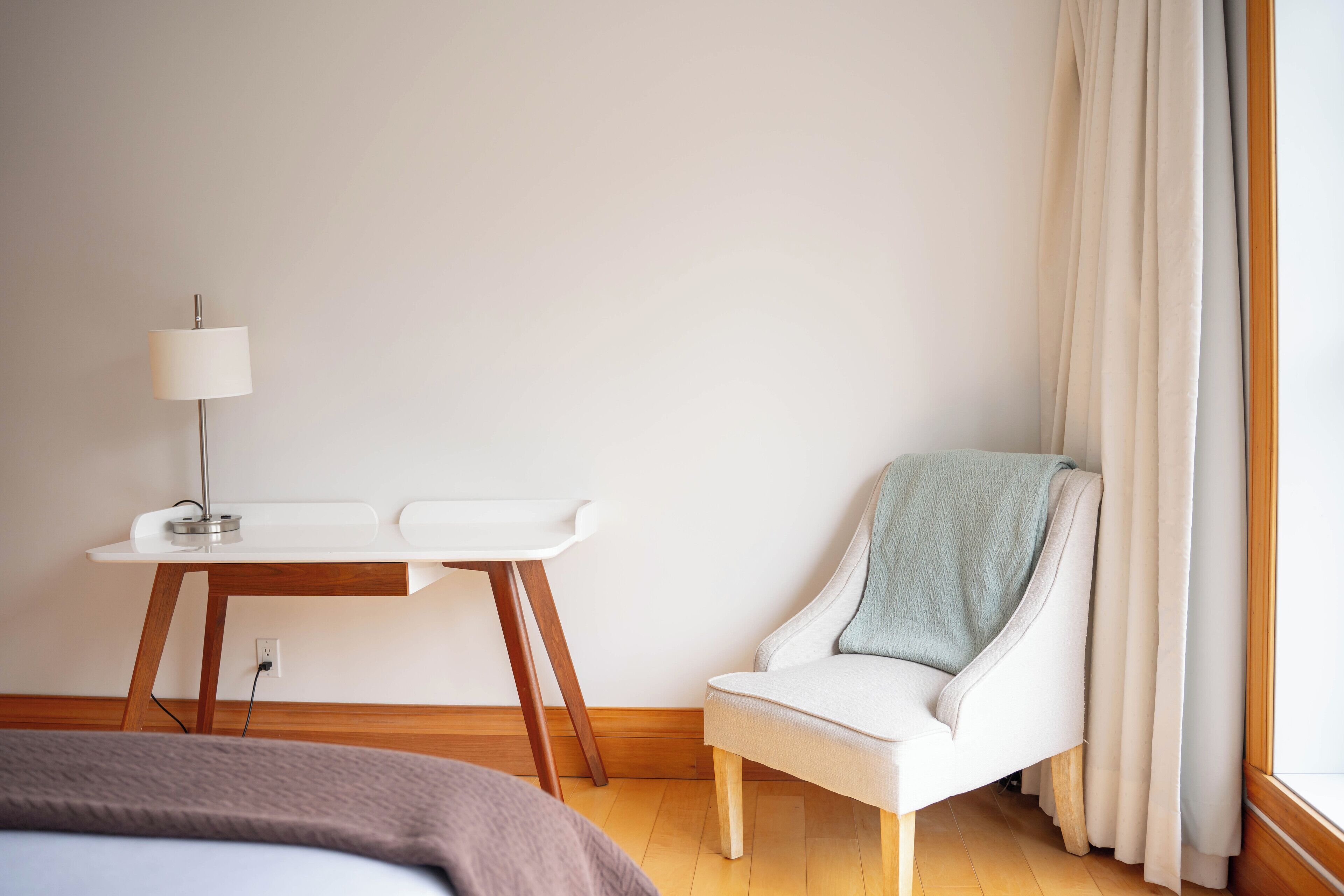 A hotel room with a bed, a desk with a lamp, a chair with a blanket, and a window with curtains.