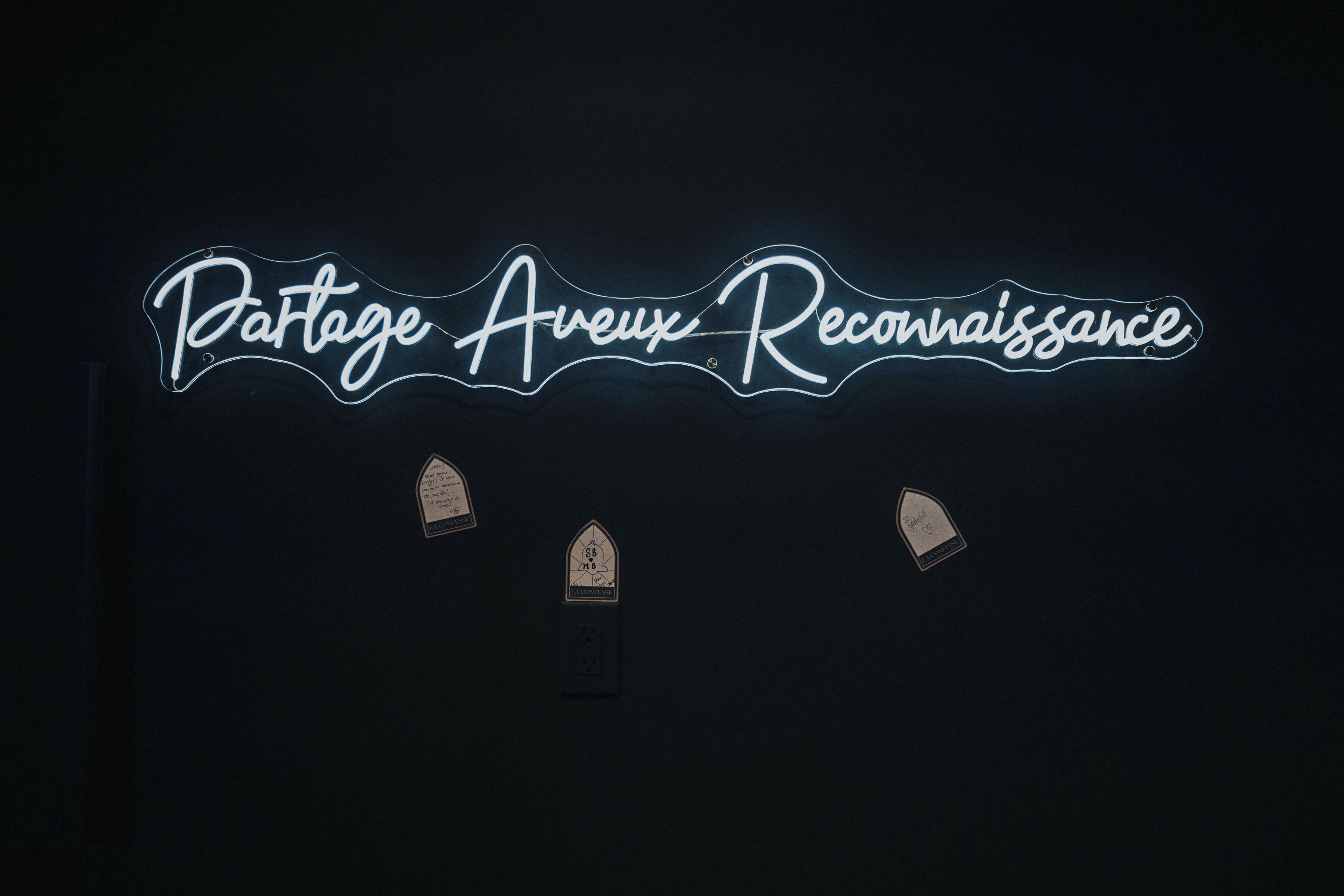 Un letrero de neón con el texto 'Partage Avenues Reconnaissance' y tres pequeños iconos con forma de casas debajo.