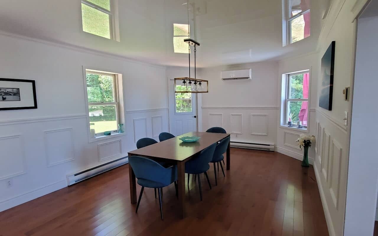 A dining area with a wooden table and blue chairs, large windows, and a hanging light fixture.