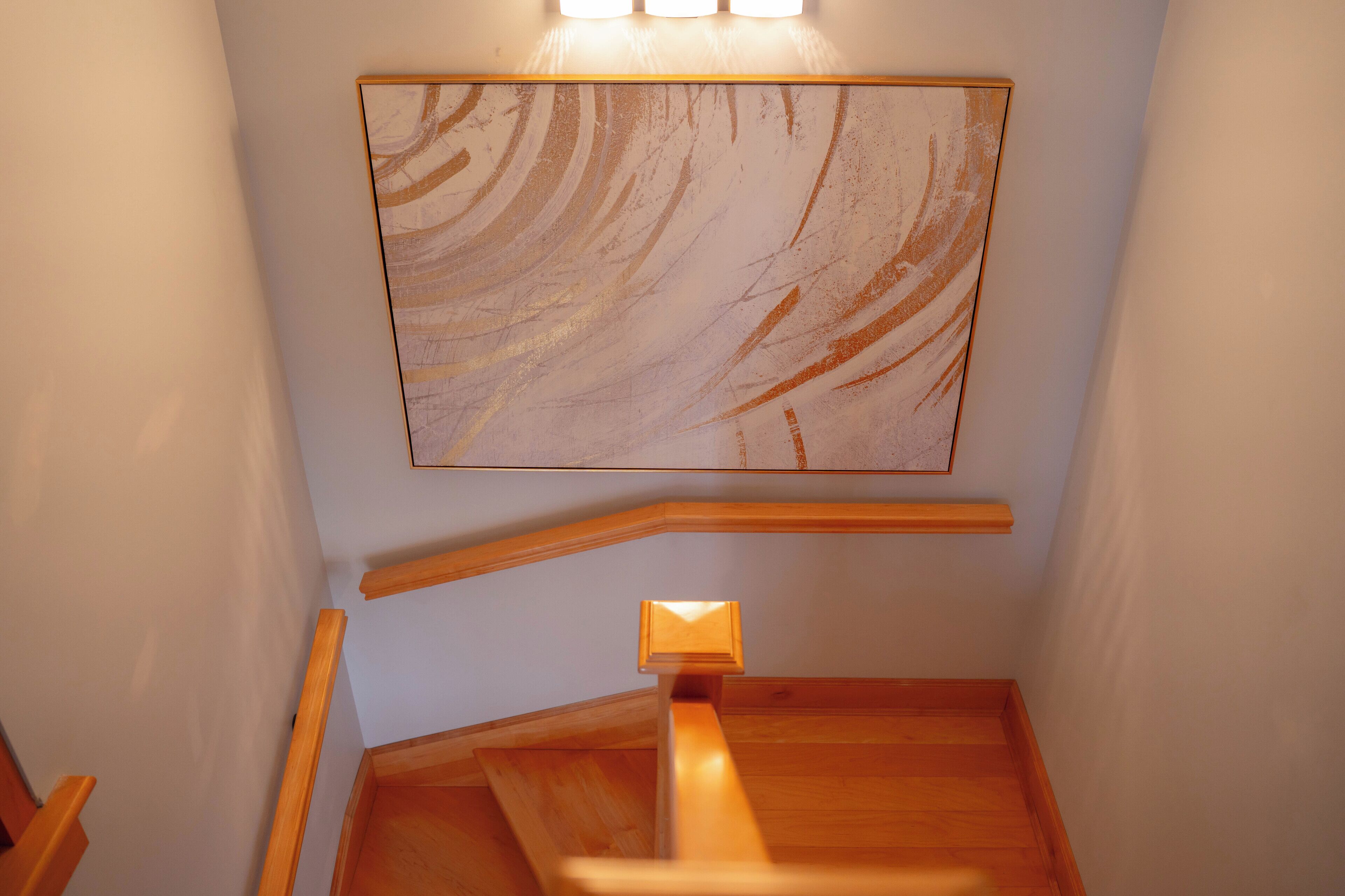 Una escalera de madera con barandilla, un aplique en la pared y un cuadro abstracto de gran tamaño.