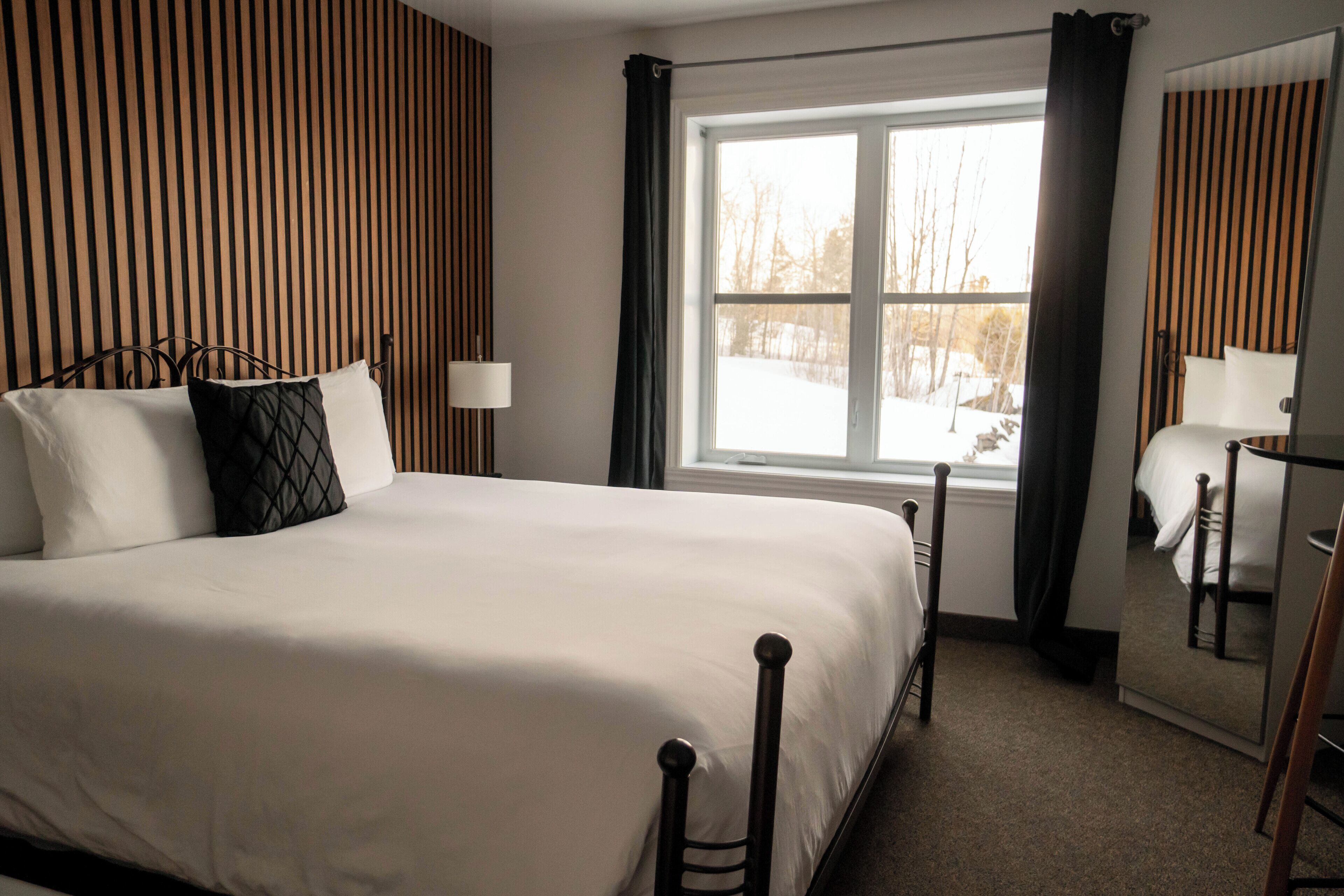A hotel room with a large bed, a window with curtains, a bedside table with a lamp, and a mirror.