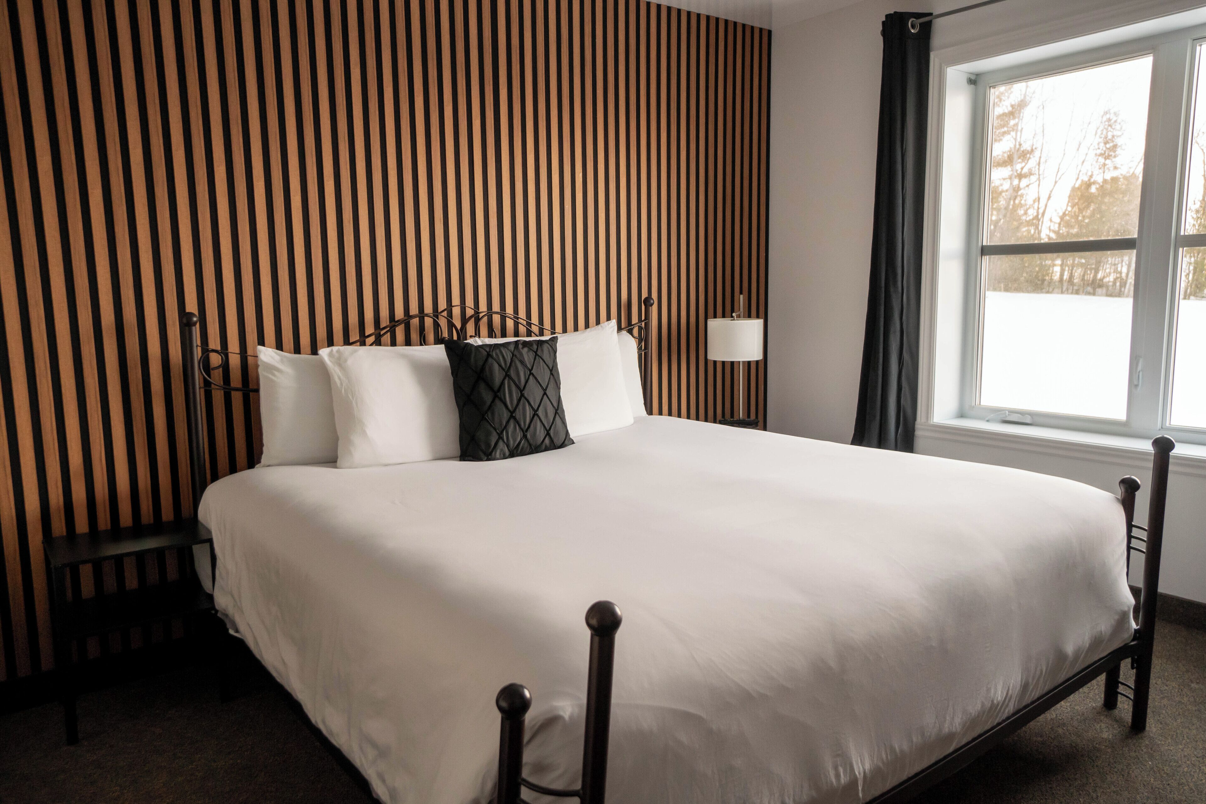 Een net opgemaakt bed in een kamer met een houten wand met strepen en een raam met gordijnen.