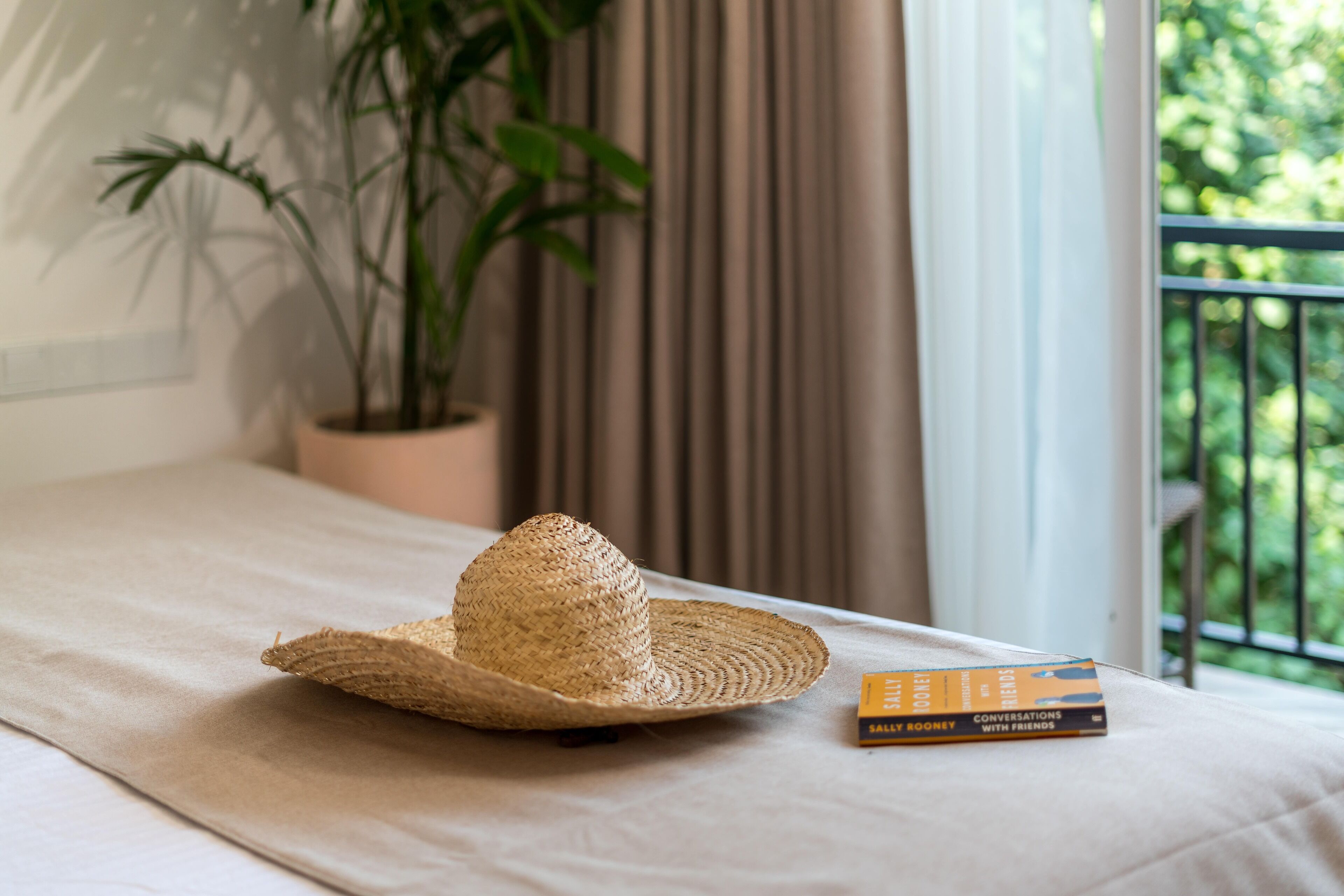 A straw hat and a book on a bed with a view of greenery outside.