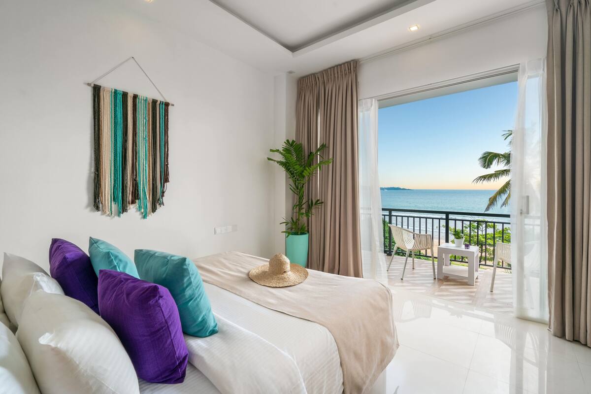 A bedroom with a bed, pillows, a plant, and a balcony with a view of the sea.