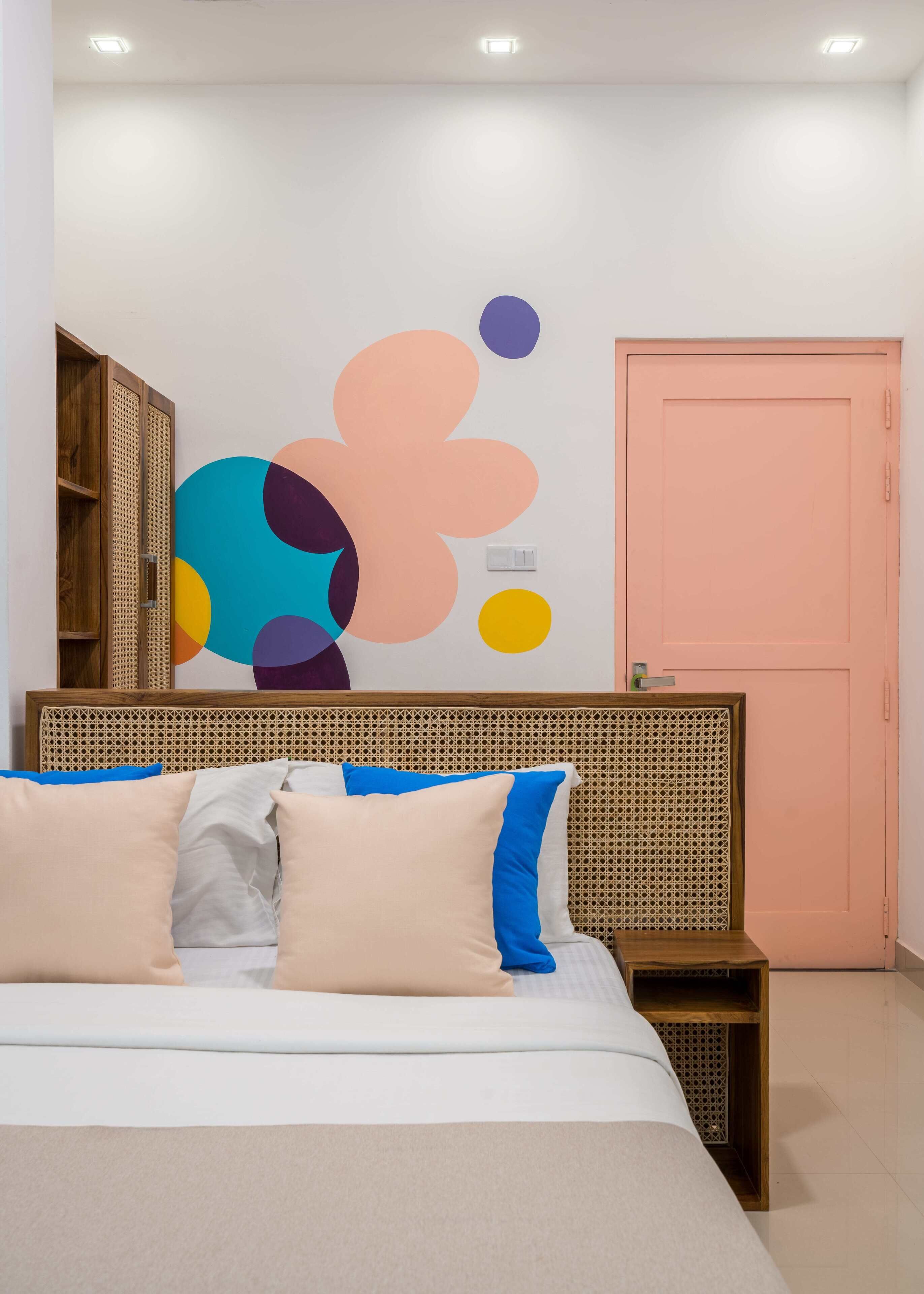 A bedroom with a bed, pillows, a rattan headboard, a wall with abstract art, a pink door, and a wooden wardrobe.