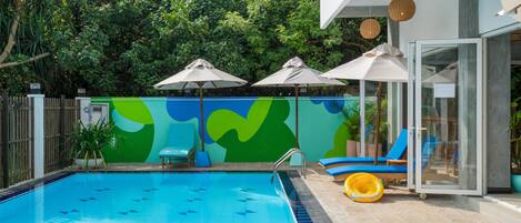 Una piscina con un mural azul y verde, sillas de descanso y sombrillas.
