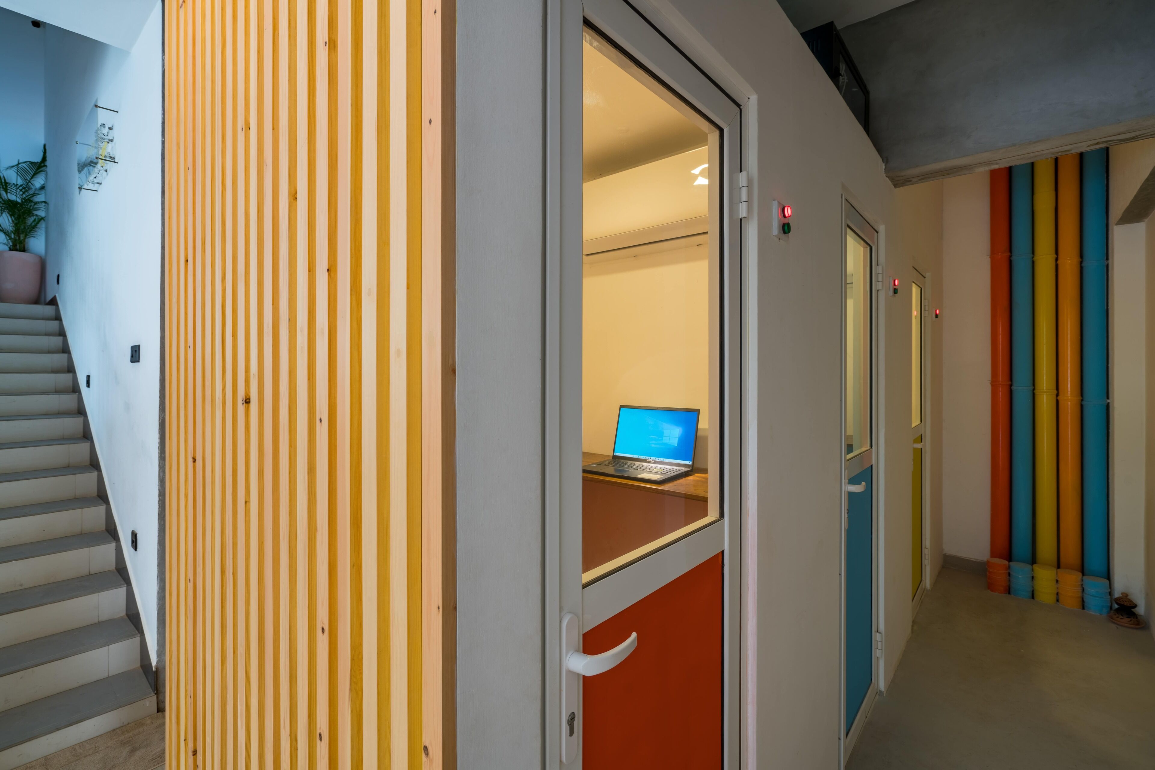 Un pasillo moderno con una escalera, una puerta y un escritorio con una computadora portátil, y elementos verticales de colores.