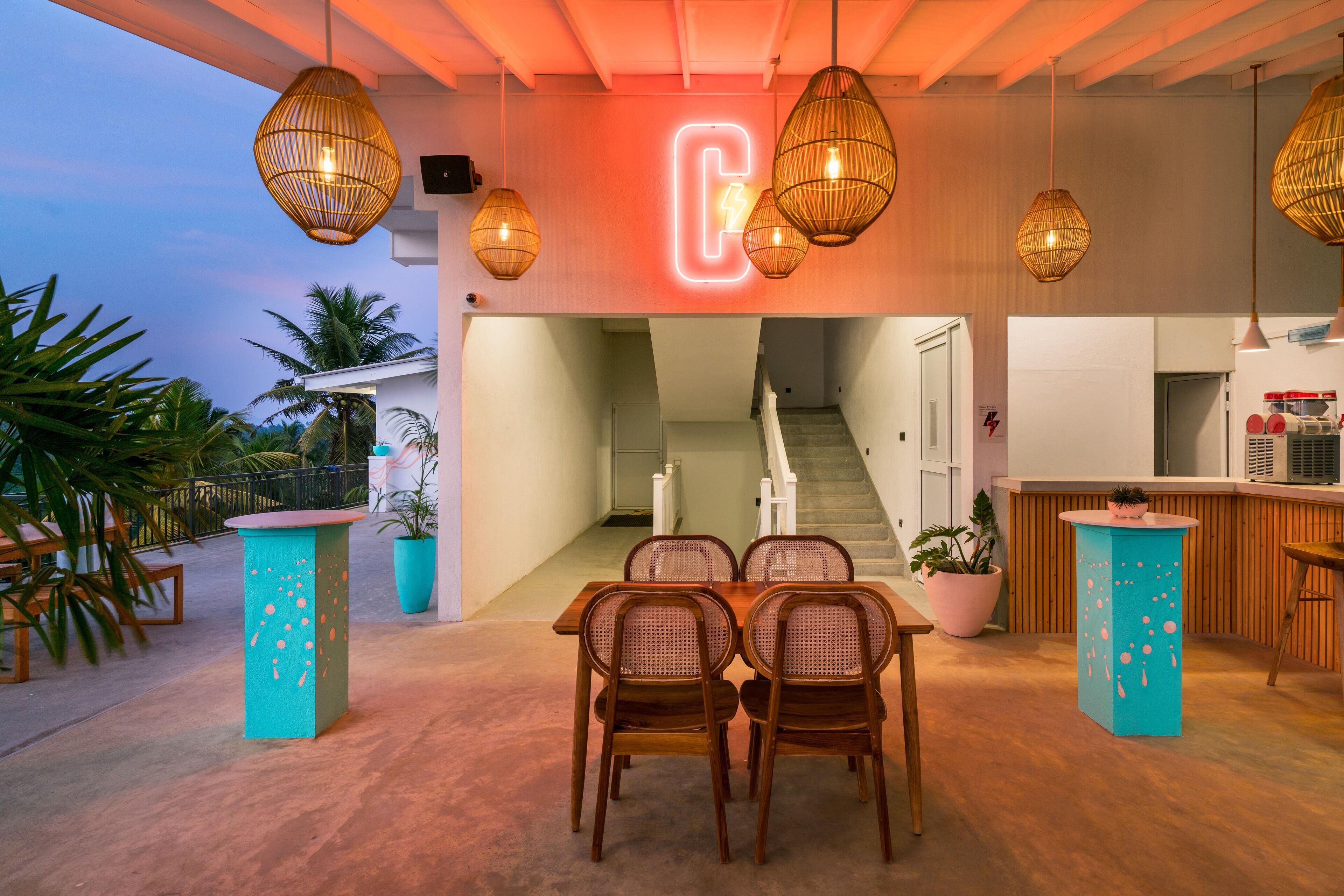 Un espacio interior moderno con un letrero de neón en forma de 'C', lámparas colgantes de mimbre, una mesa de madera con sillas y plantas en macetas.