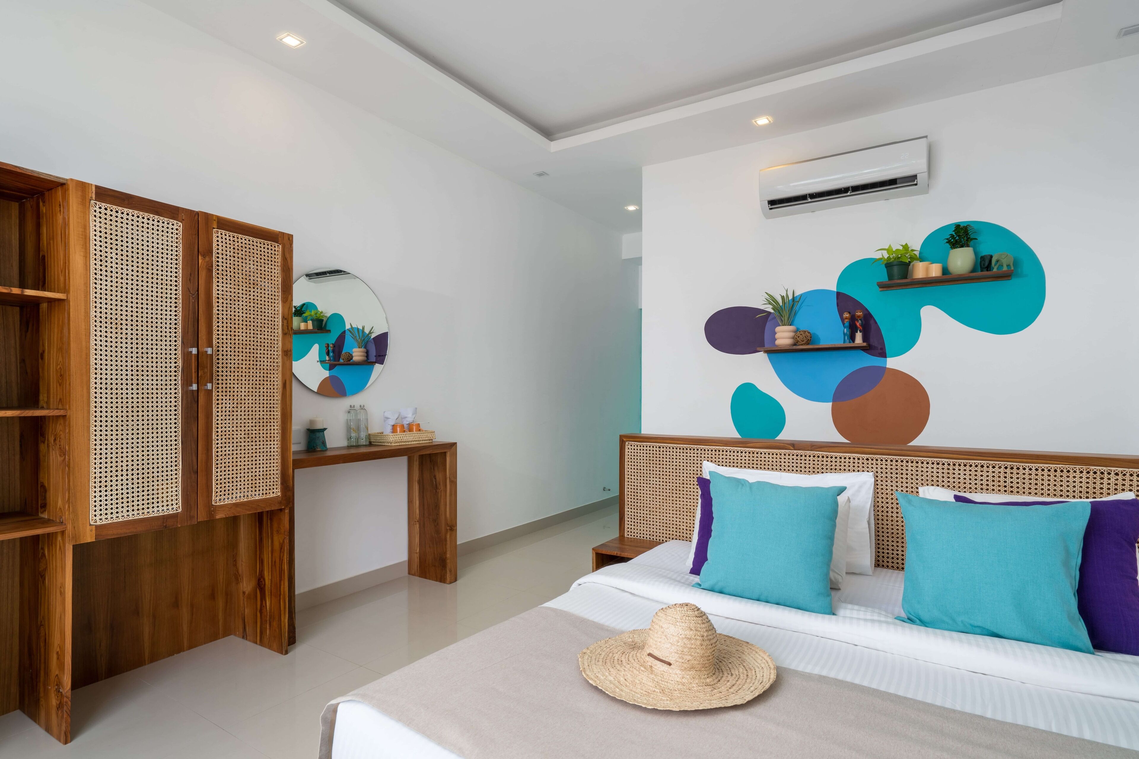 A modern hotel room with a wooden wardrobe, a bed with teal and purple pillows, a round mirror, and a wall-mounted shelf with plants.
