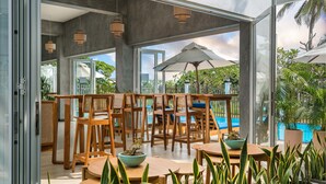 An outdoor dining area with wooden tables and chairs, surrounded by potted plants and a view of a swimming pool. - Outpost Weligama Beachfront (Weligama)