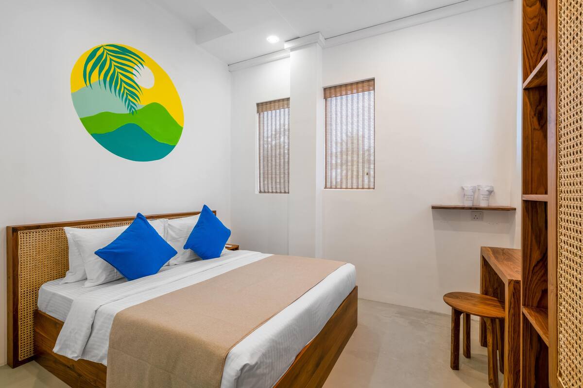 A bedroom with a bed, a wooden headboard, a desk with a chair, and two windows with blinds.
