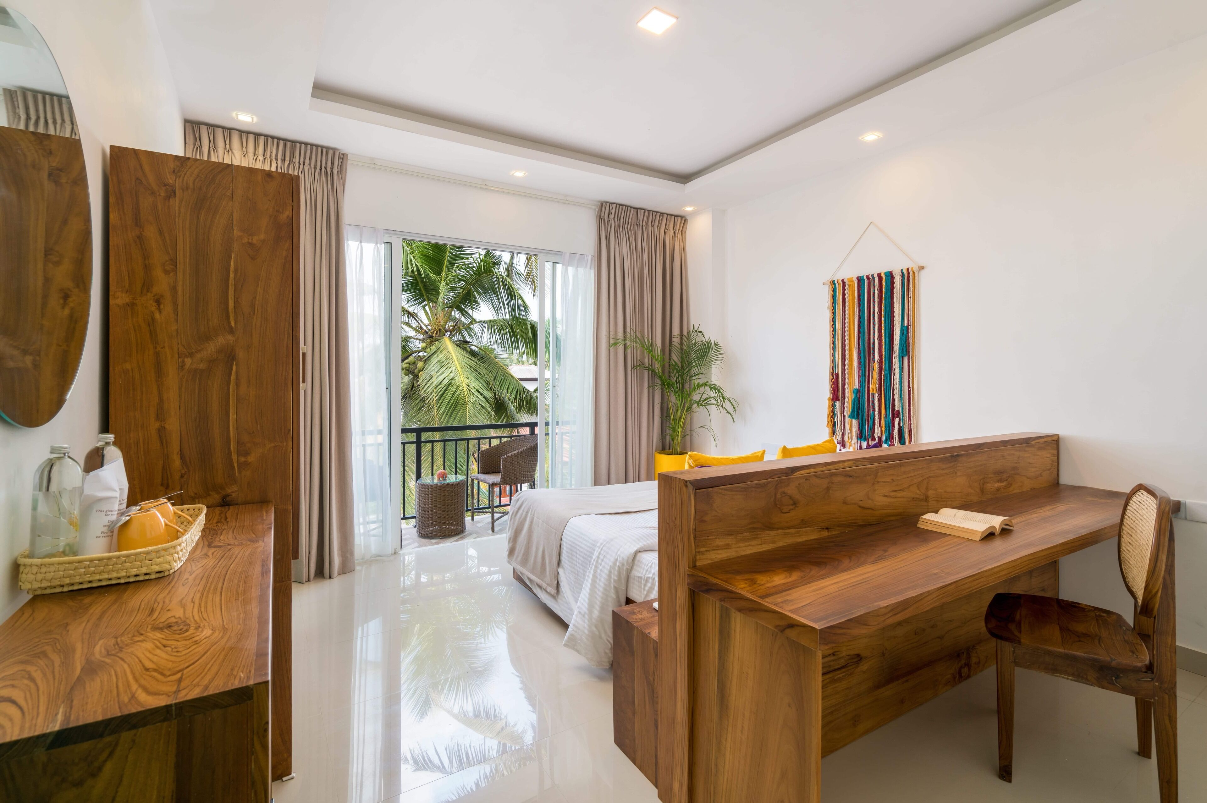 Une chambre d’hôtel moderne avec un bureau en bois, un lit recouvert de draps blancs, un balcon offrant une vue sur les palmiers et un grand miroir au mur.