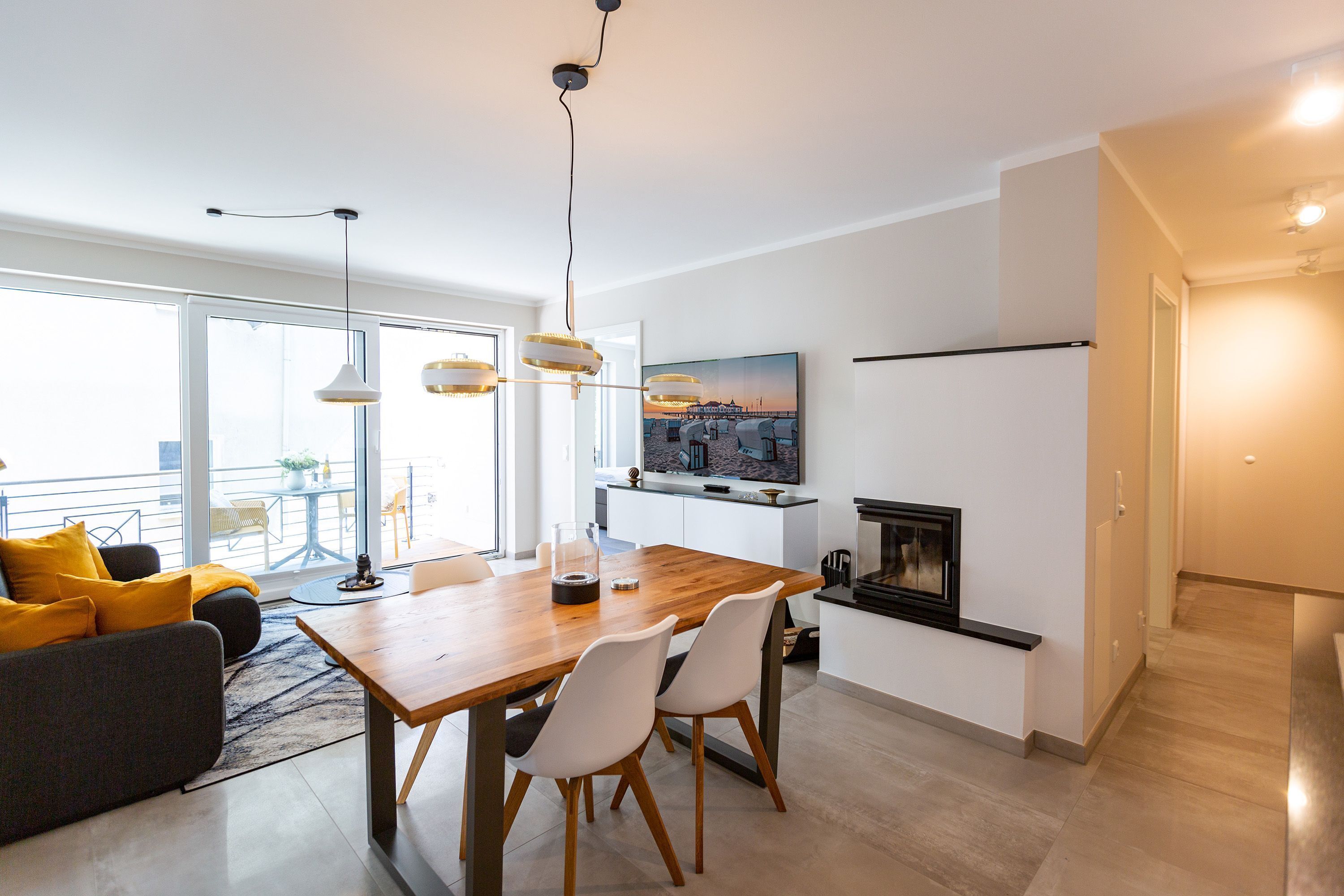 A modern living room with a wooden dining table, white chairs, and a sofa with yellow cushions. There is a fireplace and a TV mounted on the wall.