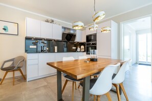 A modern kitchen with white cabinets, a wooden dining table, and white chairs. There is a large window providing natural light. - Quartier 7, App. 03, Germany (Heringsdorf)