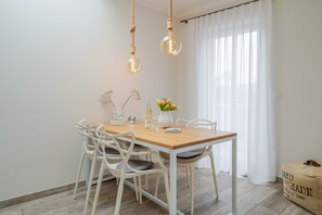 Uma área de refeições com mesa de madeira, cadeiras brancas, luminárias pendentes e uma janela com cortinas leves.