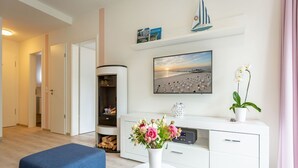 En moderne stue med en sofa, et rundt bord, et hvidt konsolbord med en vase med blomster, en reol og et maleri af en strand.