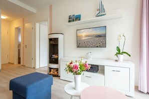 Un salon moderne comprenant un canapé, une petite table ronde, une console blanche ornée d’un vase de fleurs, une bibliothèque et un tableau représentant une scène de plage.