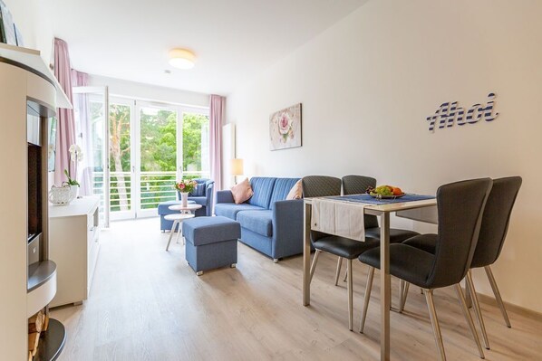 A modern living room with a sofa, dining table, and chairs. There is a large window with curtains, a painting on the wall, and a TV on the left.