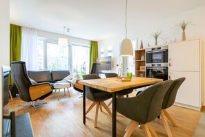 Un salon moderne avec une table à manger en bois, des chaises noires et grises, un canapé et un coin cuisine équipé d’un four et de placards.
