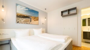 Una cama doble con ropa de cama blanca, una mesita de noche, un estante en la pared, un cuadro con una playa y un baño con espejo y lavabo.