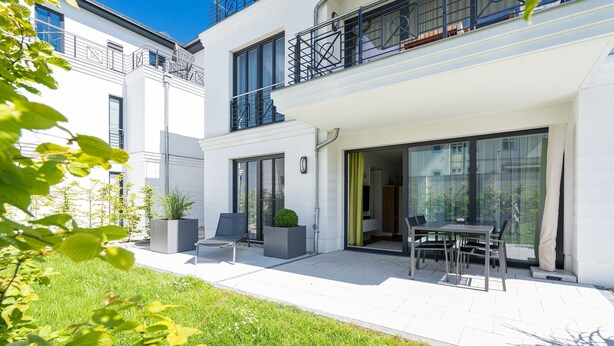 Un edificio moderno de dos pisos con un patio que cuenta con una mesa, sillas, un pequeño jardín y una entrada con puerta de vidrio.