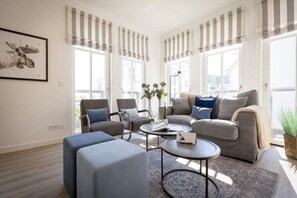 Una sala moderna con un sofá gris, un sillón azul y una mesa de centro redonda. Hay cortinas a rayas en las ventanas y un cuadro en la pared.